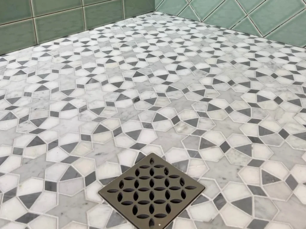 Wet room installation with green wall tiles, patterned marble floor tiles and a square drain cover.